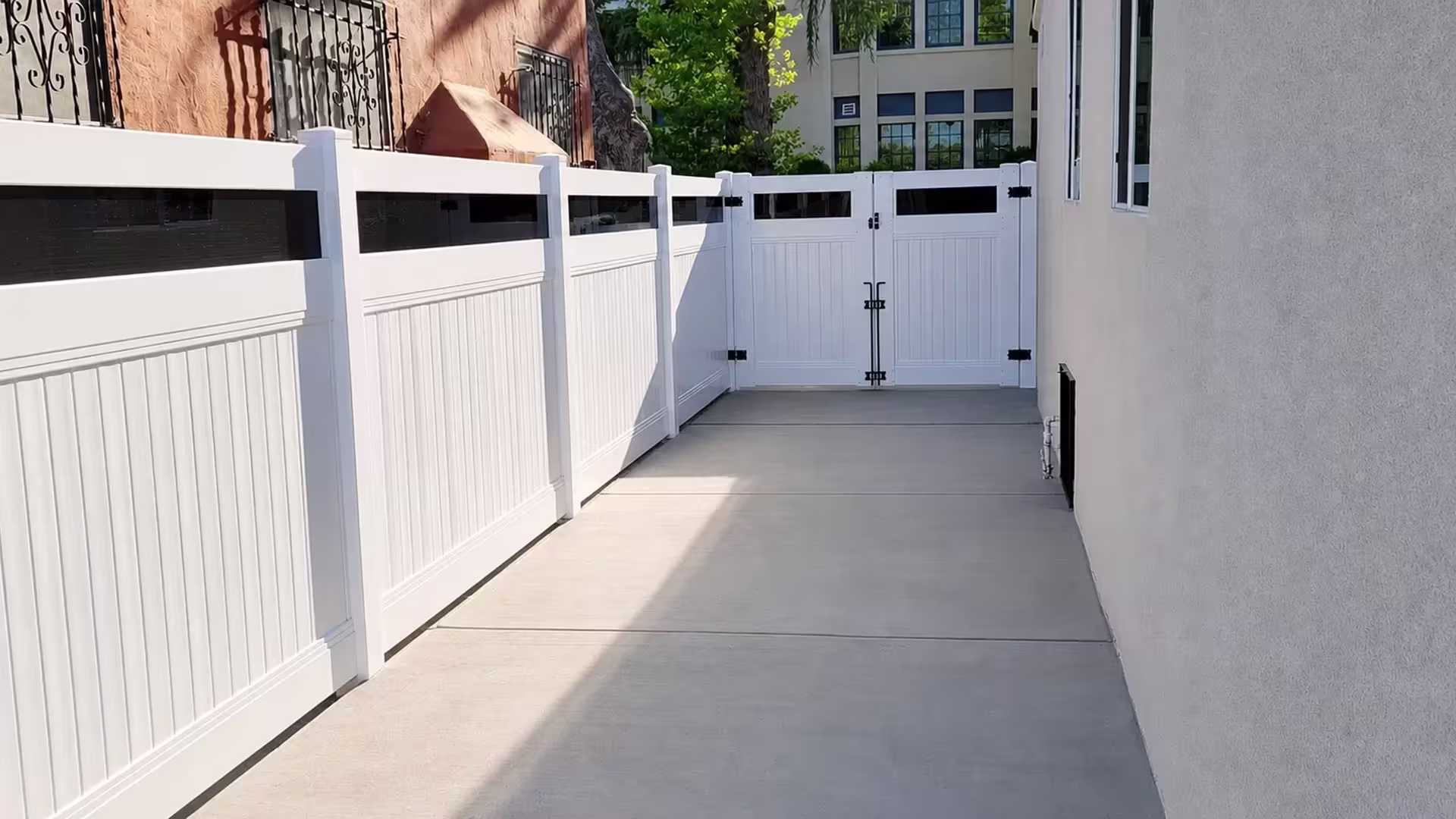 Iron window grilles mark the stucco walls beside this White vinyl privacy fence with acrylic ending at a two leaf gate