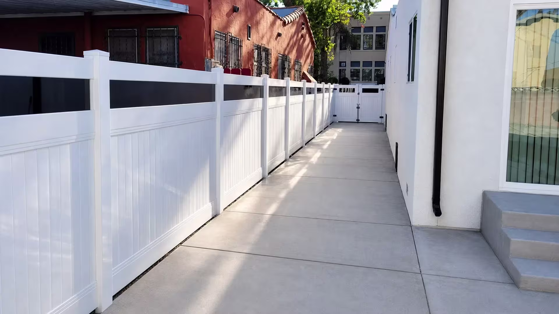 Two neighboring buildings frame this White vinyl privacy fence with acrylic threading toward a distant two leaf gate