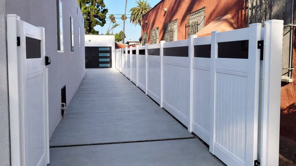 White vinyl privacy fence with acrylic and matching gates flank both sides of the driveway near shrubs and trees