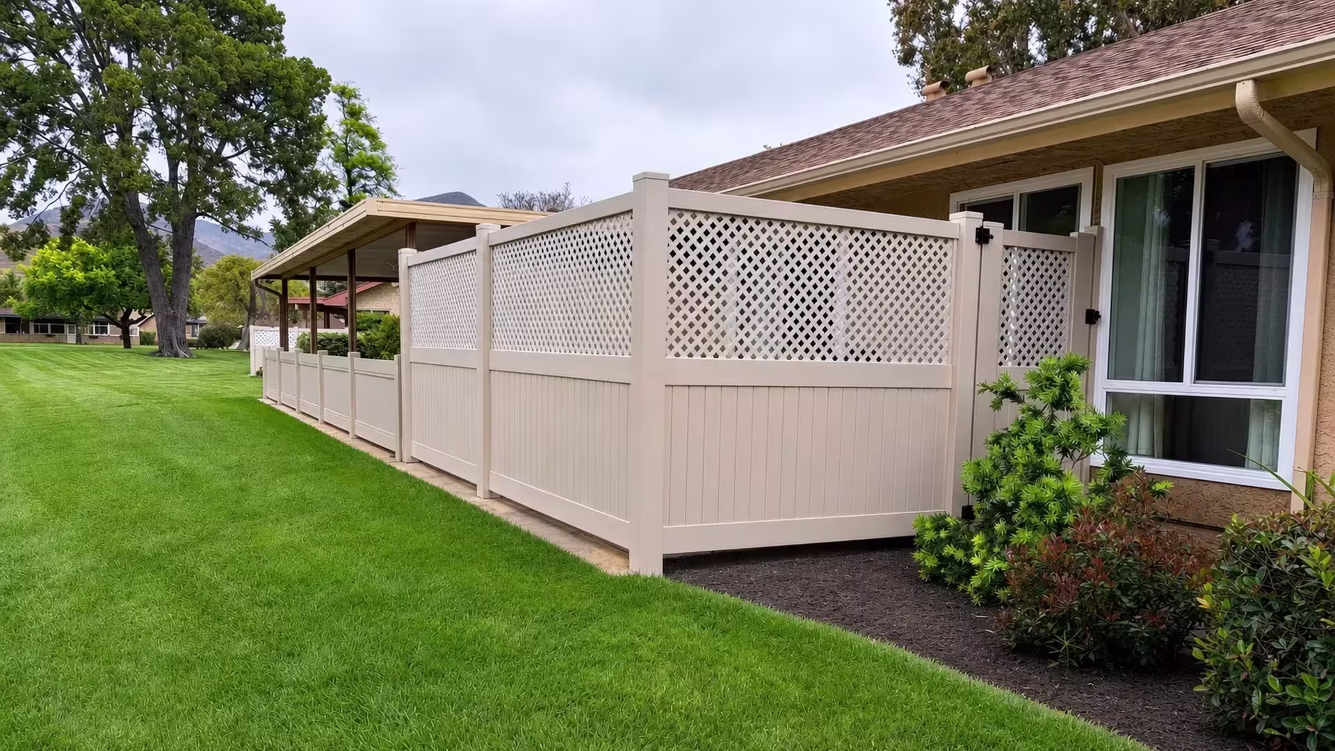Tan vinyl privacy fence with a lattice top stepping across the lawn to a narrow gate beside the house
