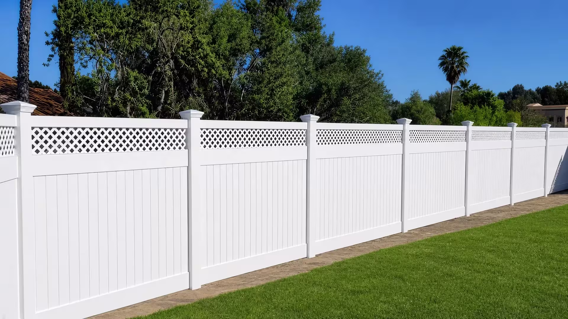 White vinyl privacy fence receding across a lawn past a stone paver strip and distant palms