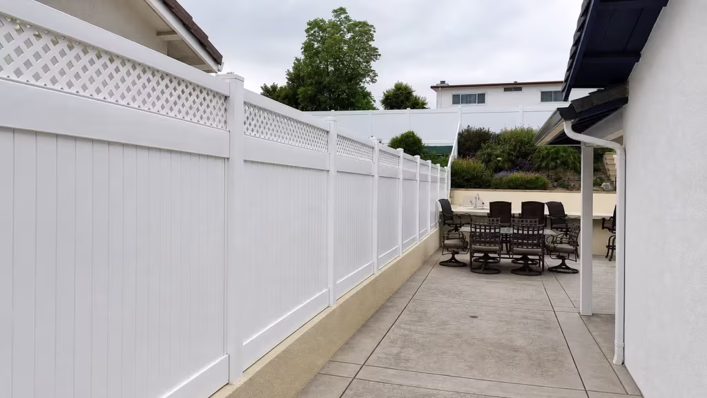 A concrete patio edge borders White vinyl privacy fence with lattice panels beside stucco house walls