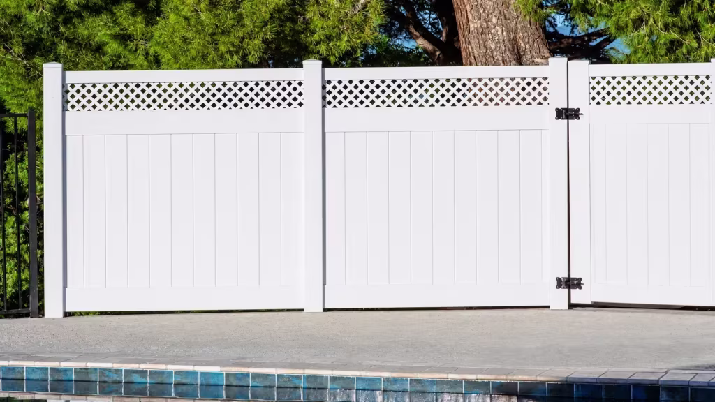 Mature pine trees stand behind White vinyl privacy fence and a matching lattice topped gate beside a blue tiled pool edge