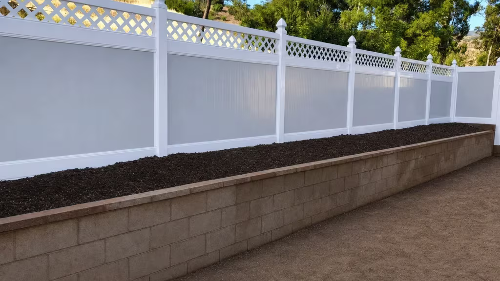 Stepped panels of White vinyl fence follow a sloped hillside grade beside a masonry retaining wall