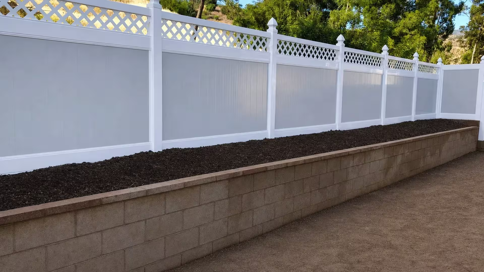 Stepped panels of White vinyl fence follow a sloped hillside grade beside a masonry retaining wall