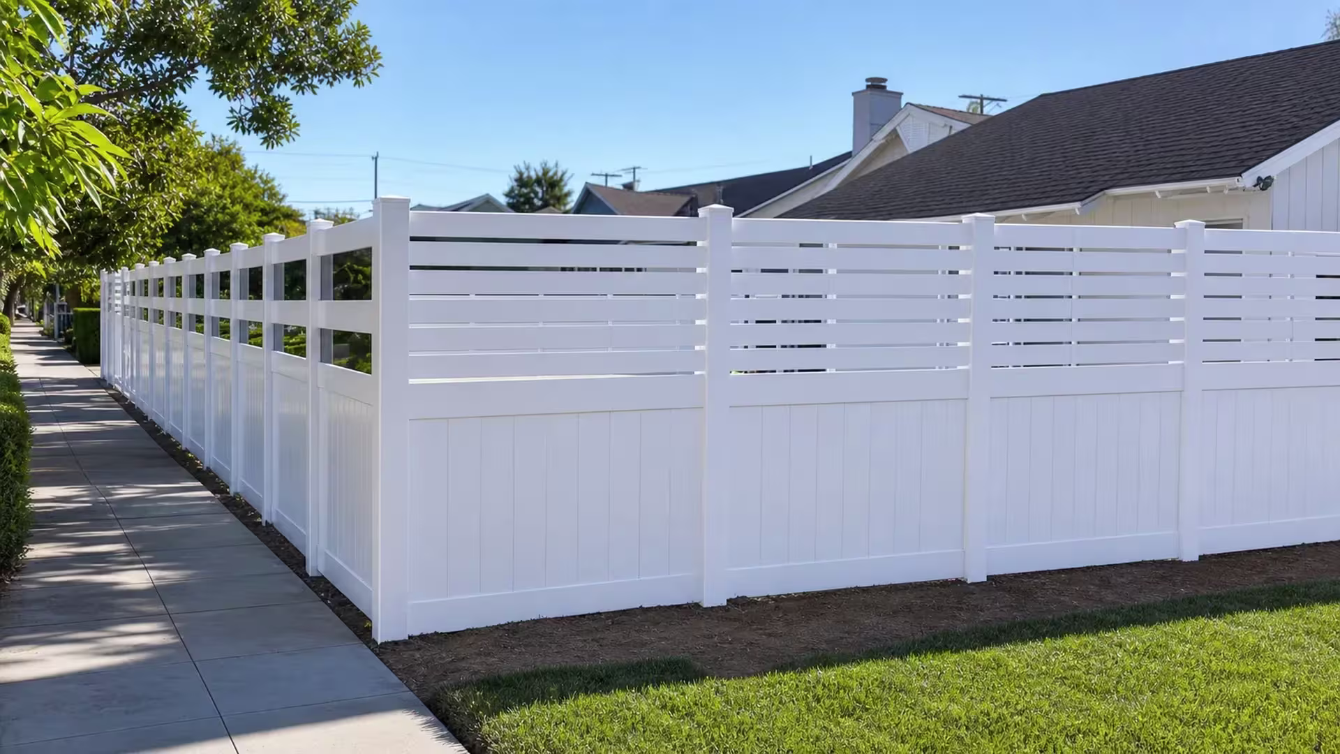White vinyl privacy fence with a picket top turning the corner and receding along a sidewalk under neighboring rooflines