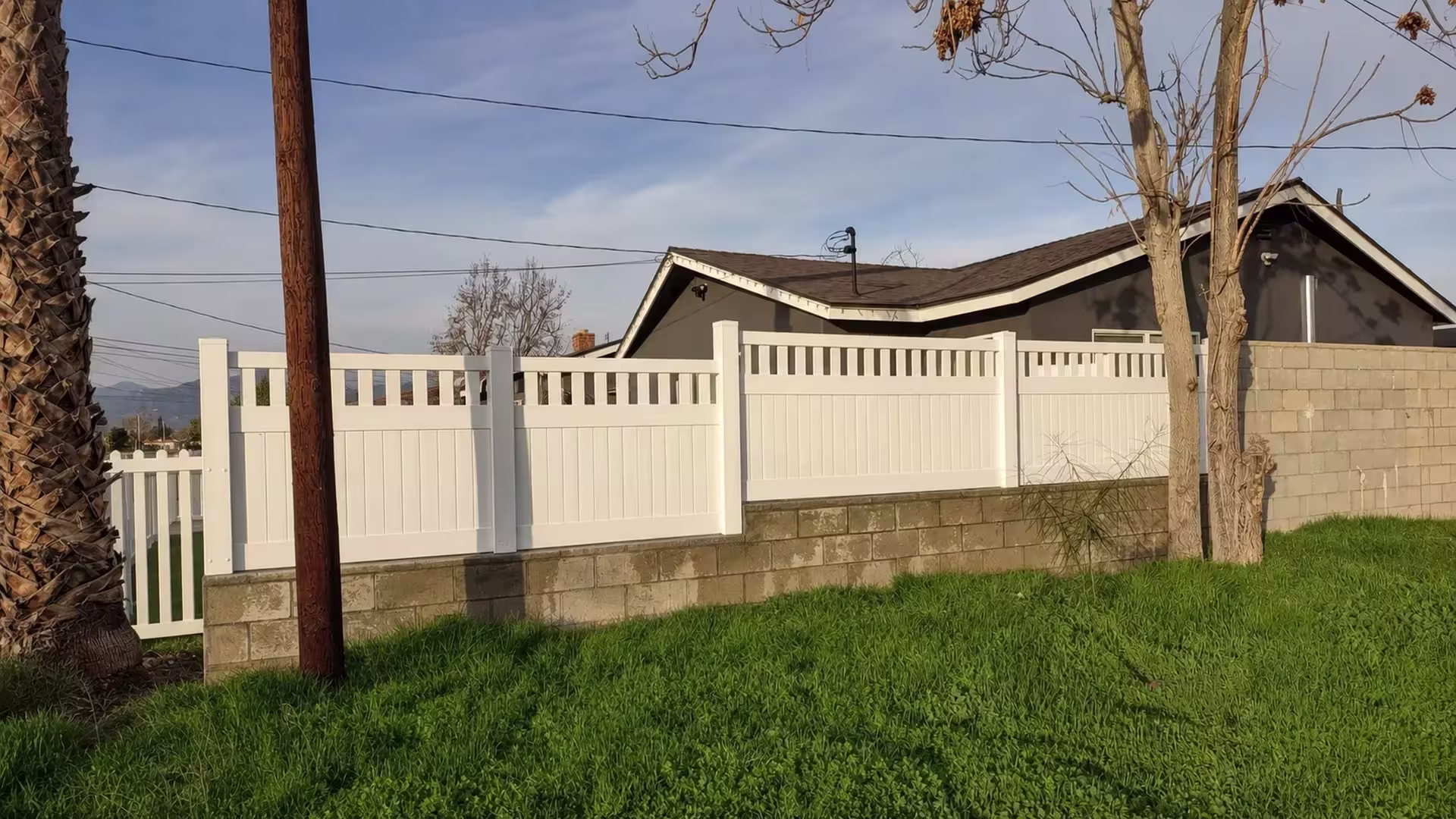 Set atop a tan concrete block retaining wall, this White vinyl fence adds a picket top near a utility pole