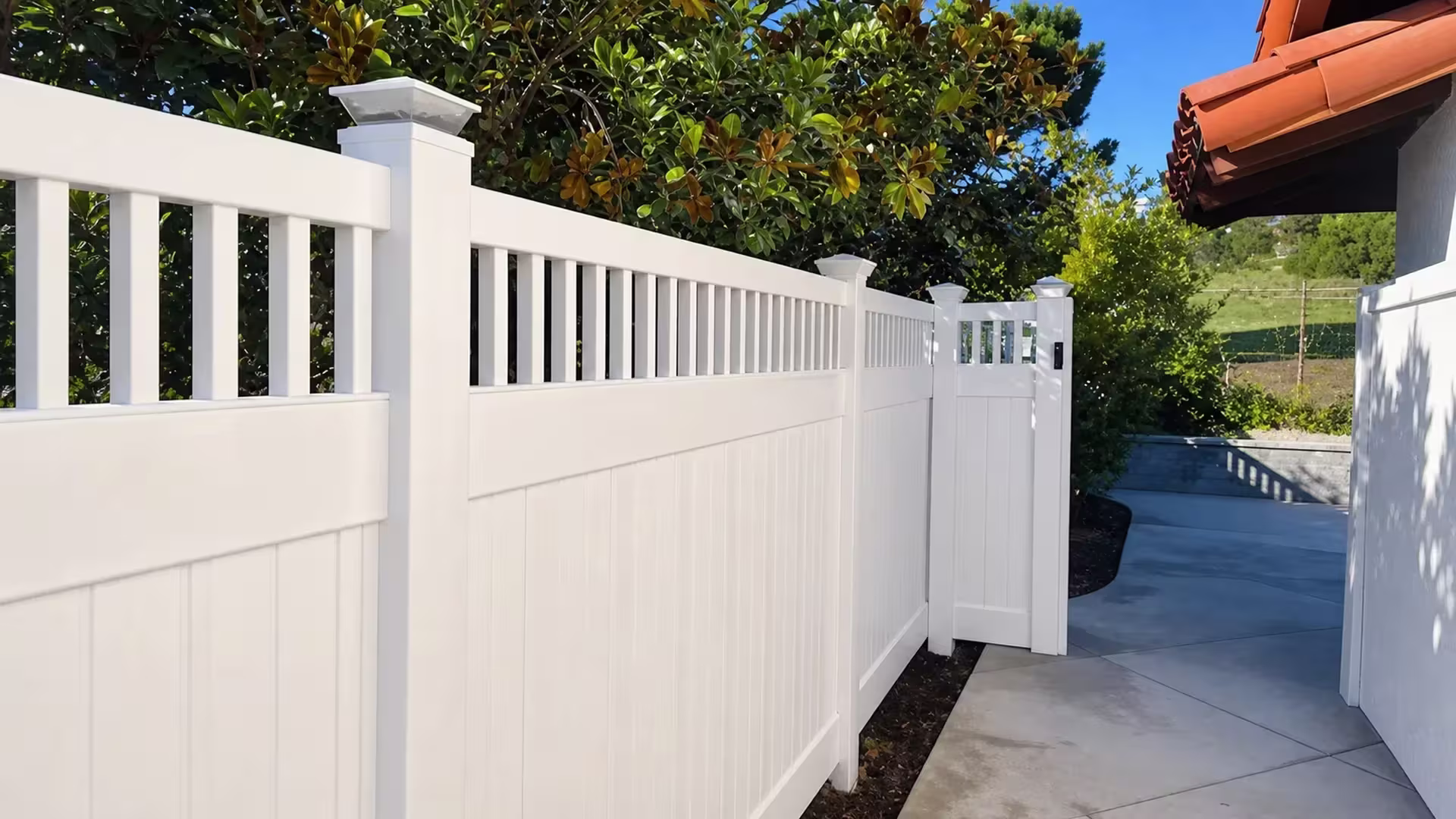 A narrow soil bed and the house eaves border this White vinyl fence, its picket top left open above