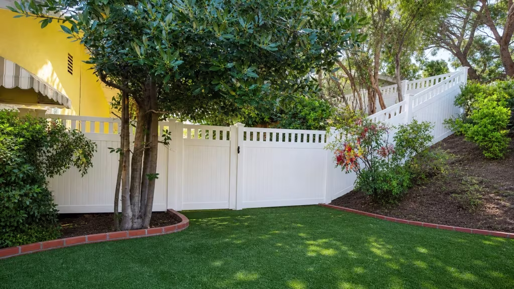 White vinyl privacy fence wraps a multi-trunk tree before its picket top climbs a planted slope