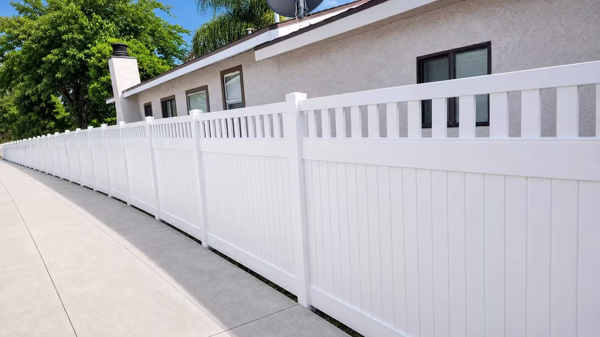 Running diagonally beside a stucco house and chimney, this White vinyl fence carries a picket top