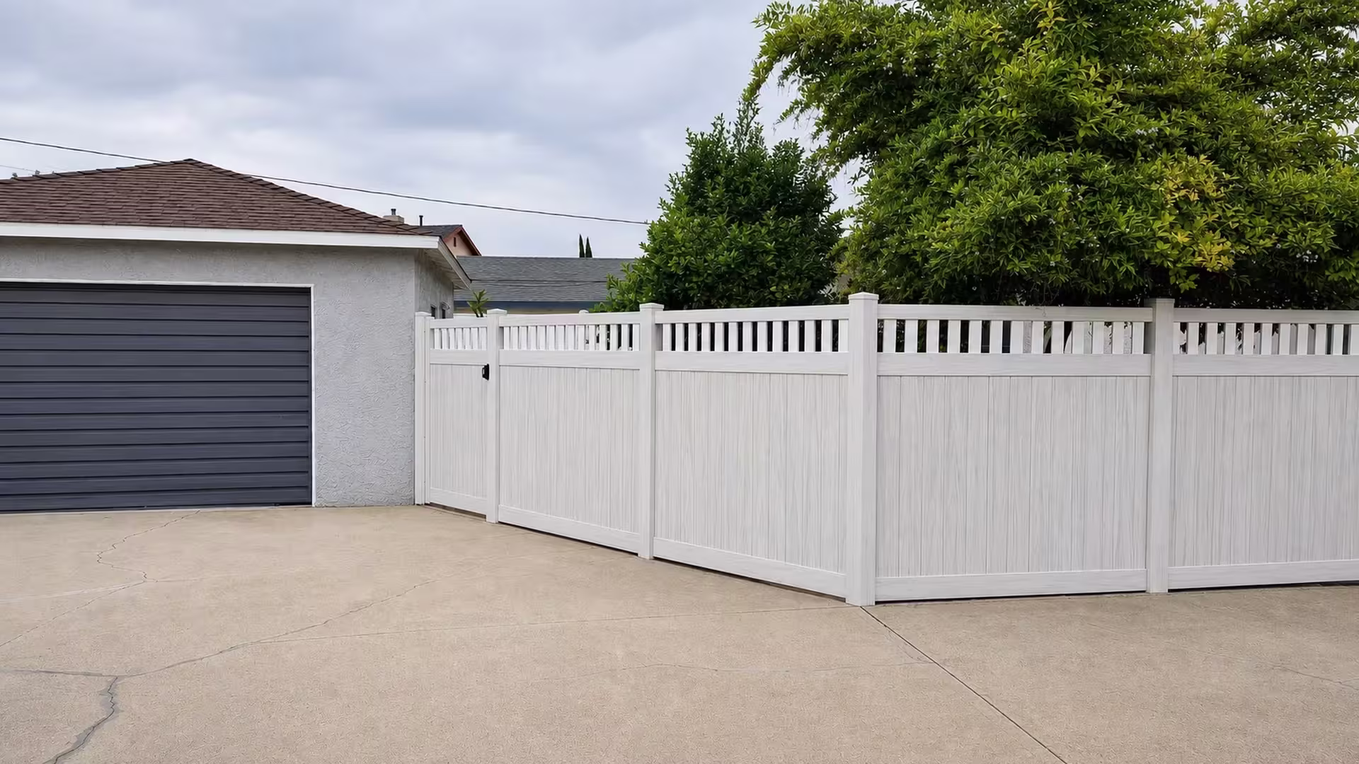 Cracked concrete paving fronts a dark sectional garage door beside this White vinyl fence where the picket top meets the gate