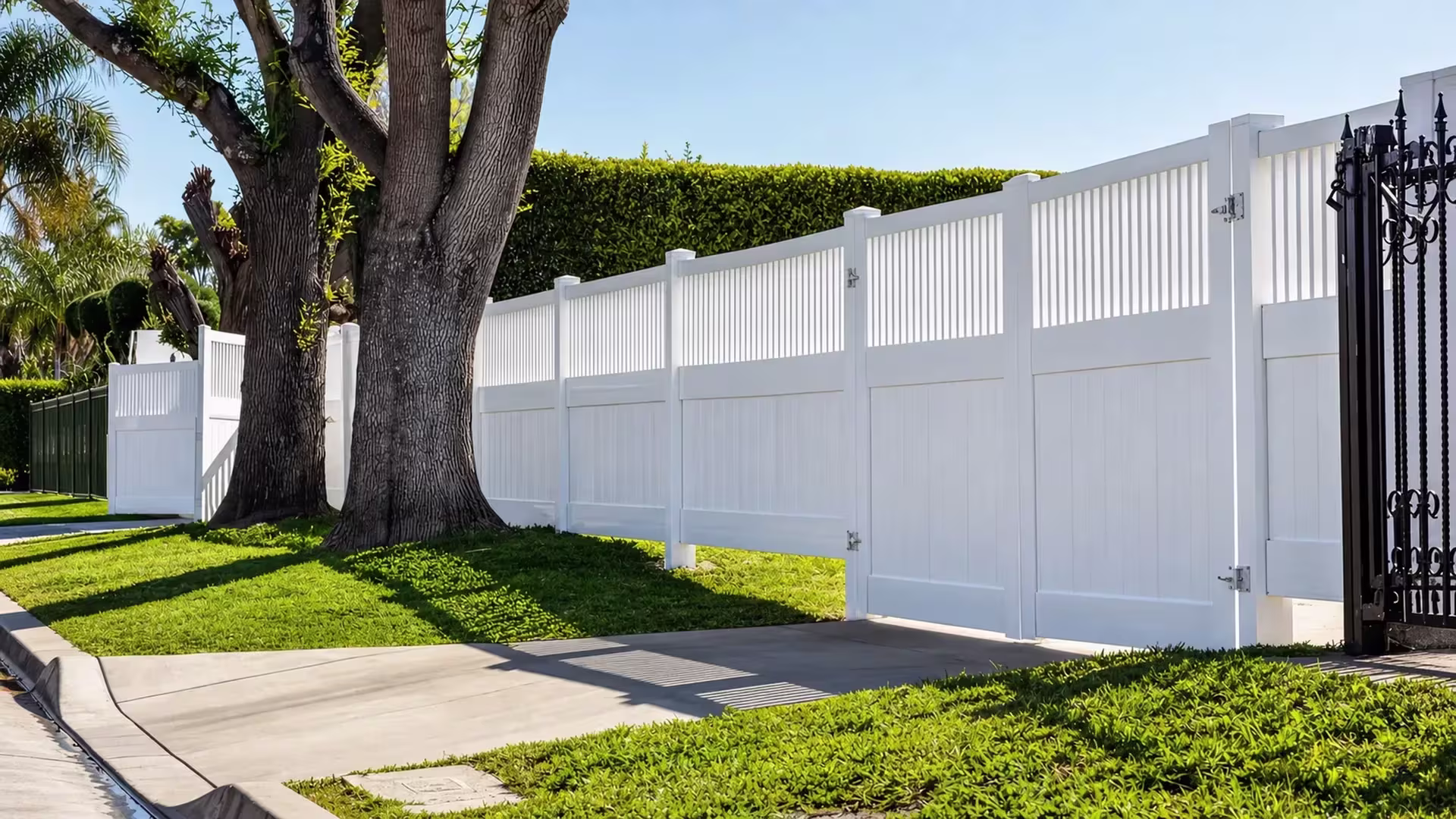 Stepped White vinyl fence panels climb the sloped yard beside a dark iron gate and hedge, each topped with pickets