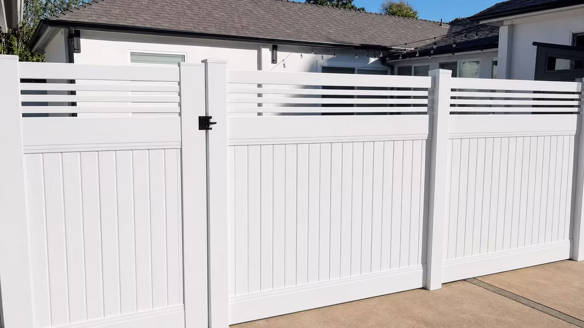 White vinyl privacy fence and gate share a picket top, the wide leaf fitted with a black latch and hinge