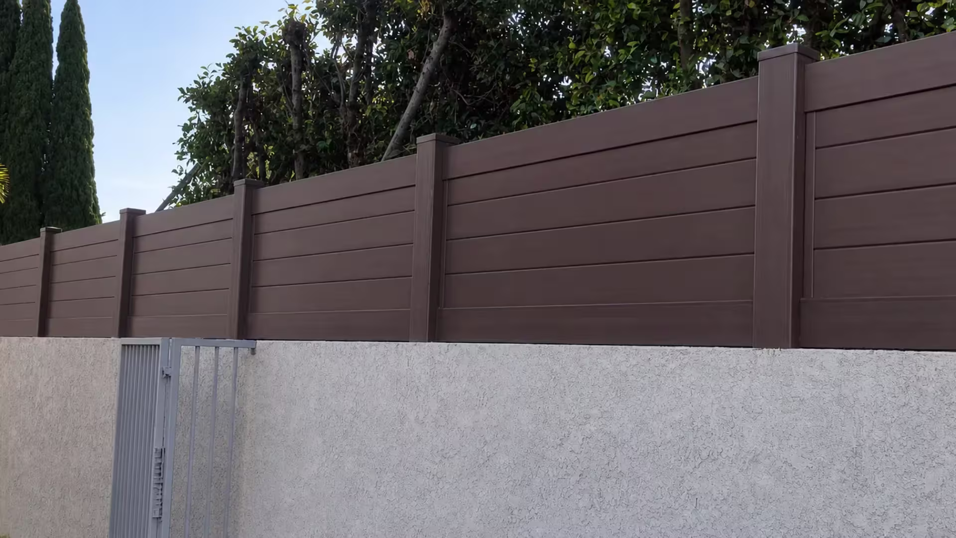 Dark Walnut vinyl wall topper capping a stucco boundary wall near a gray metal utility gate and cypress trees