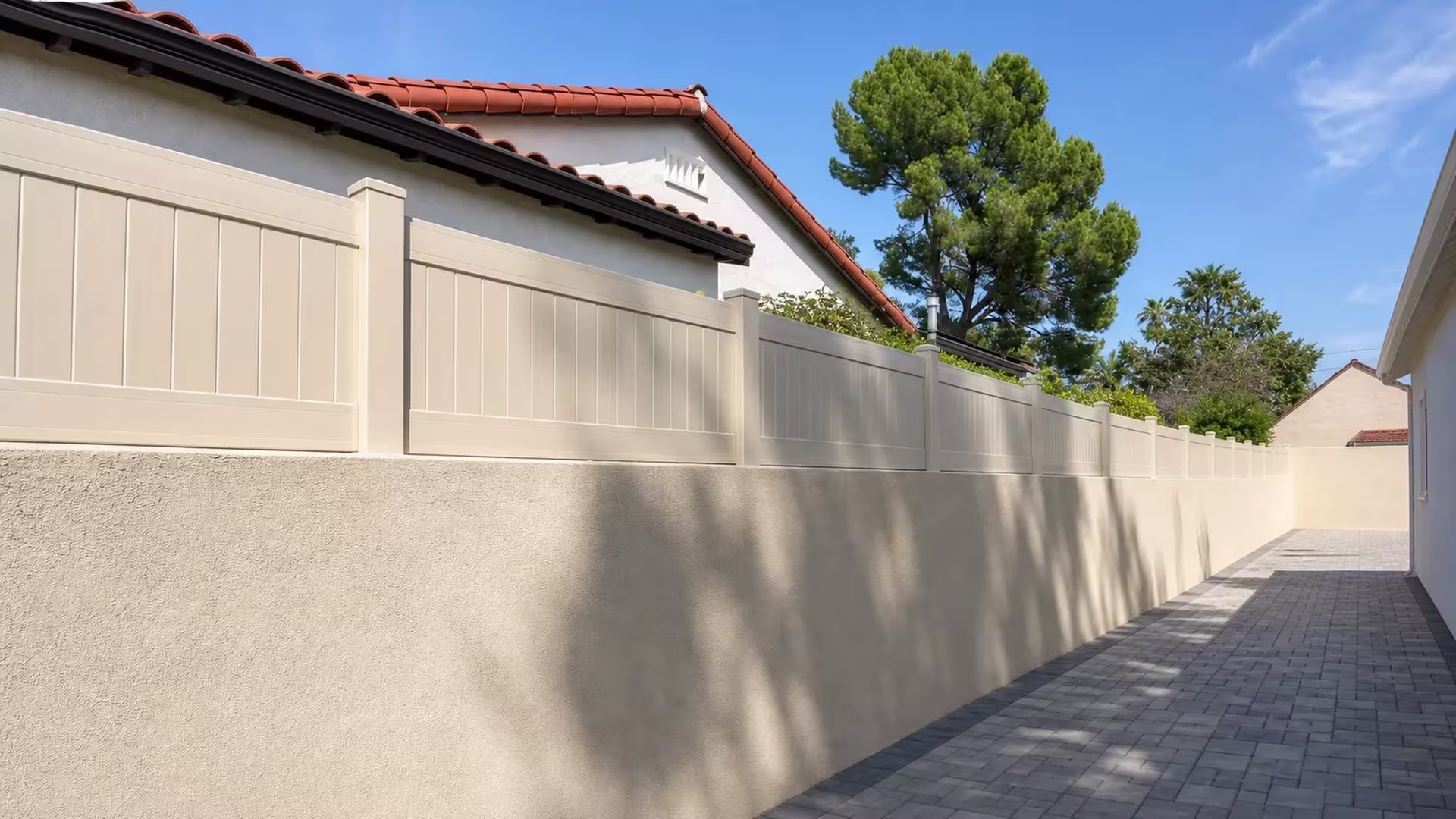 Tan vinyl privacy wall topper recedes along a beige stucco wall past a gray paver walkway and tile roof house