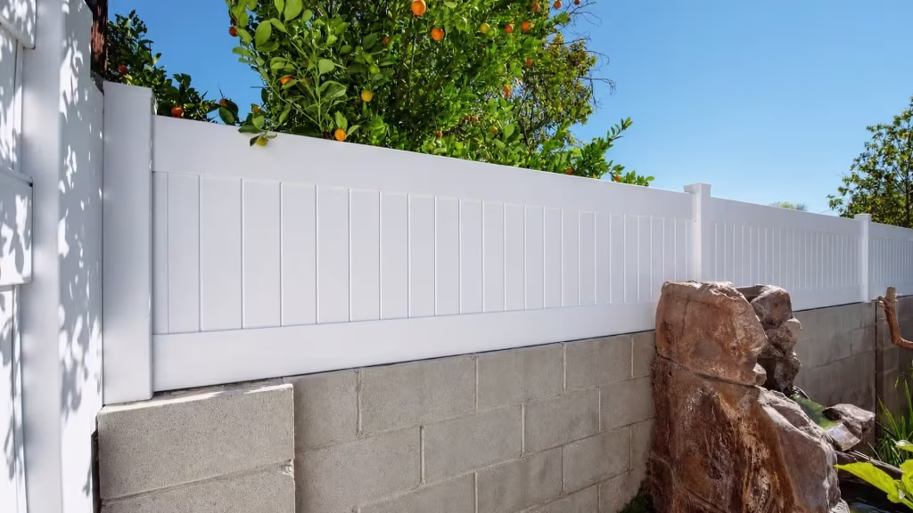 White vinyl privacy topper steps down a block wall beside a rock water feature and fruiting greenery