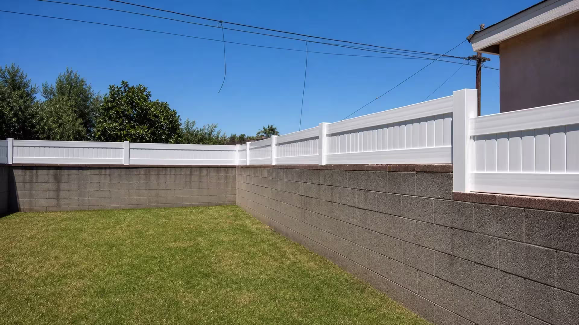 Overhead utility wires and a roof edge frame a White vinyl privacy wall topper on the graded retaining wall