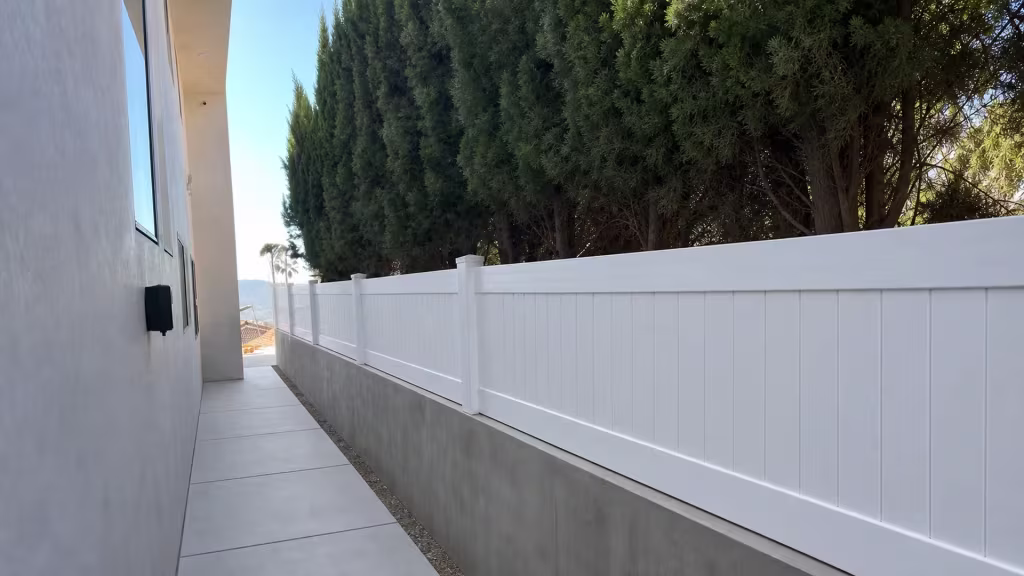 Narrow passage between a gray house wall and dense evergreen trees holds a White vinyl privacy wall topper