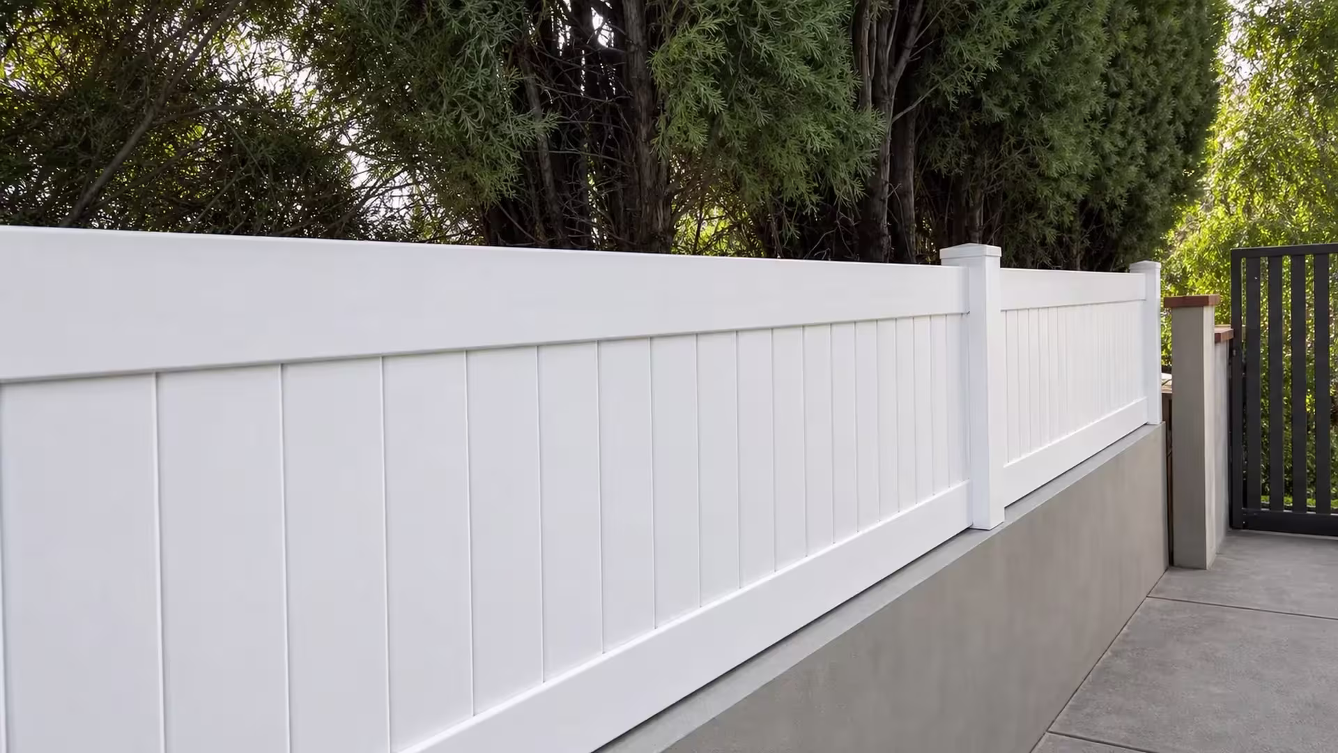 Dark metal gate and masonry posts mark the end of a White vinyl privacy wall topper run