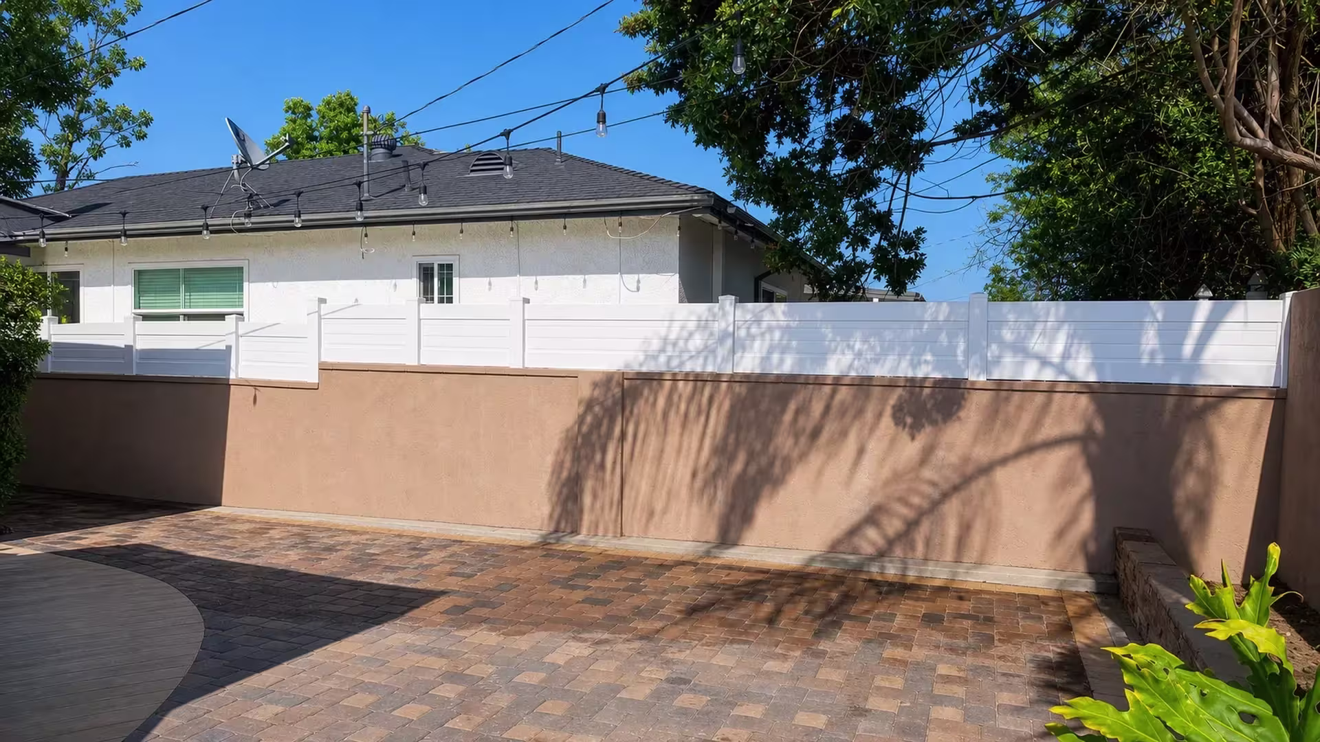 Brick paver patio and string lights sit below a White vinyl horizontal-board privacy wall topper on the tan stucco wall