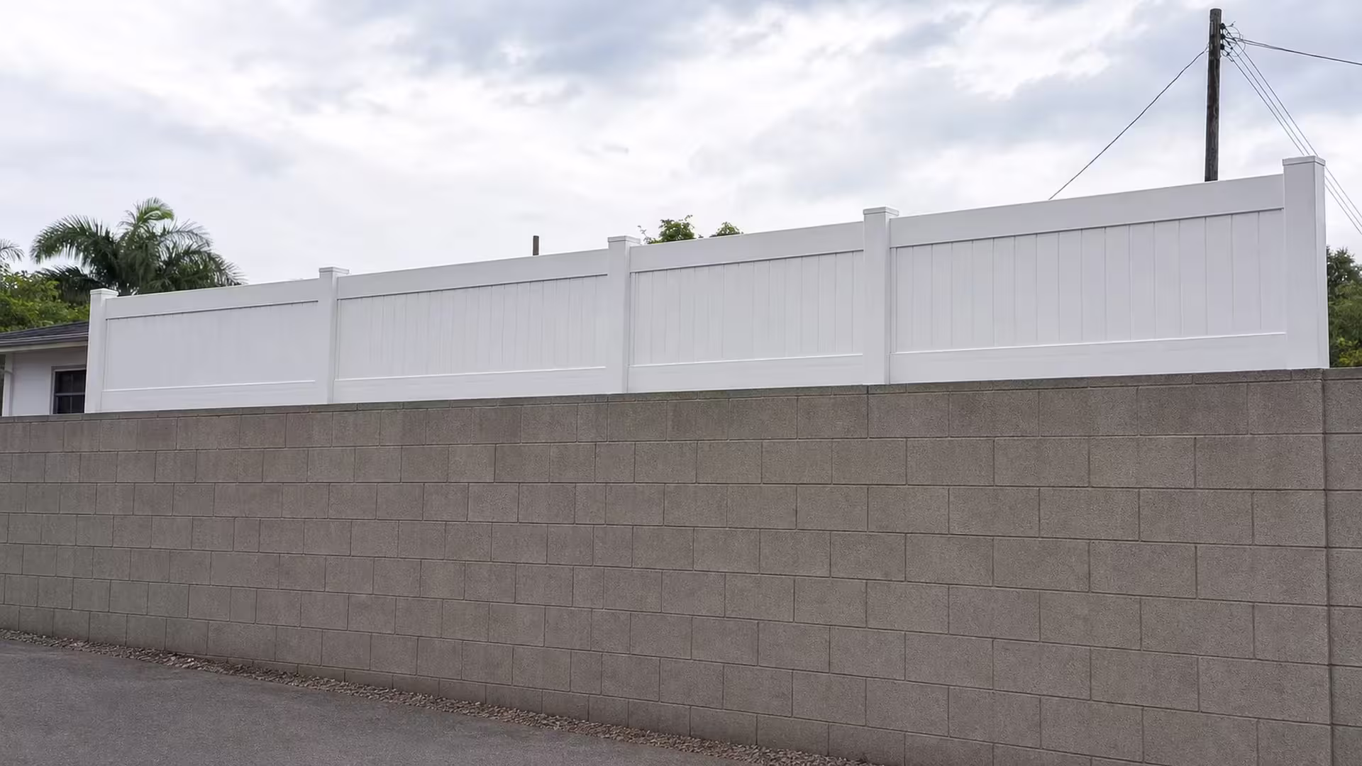 Palm fronds and overhead utility wires overhang a White vinyl privacy topper rising with the graded block wall