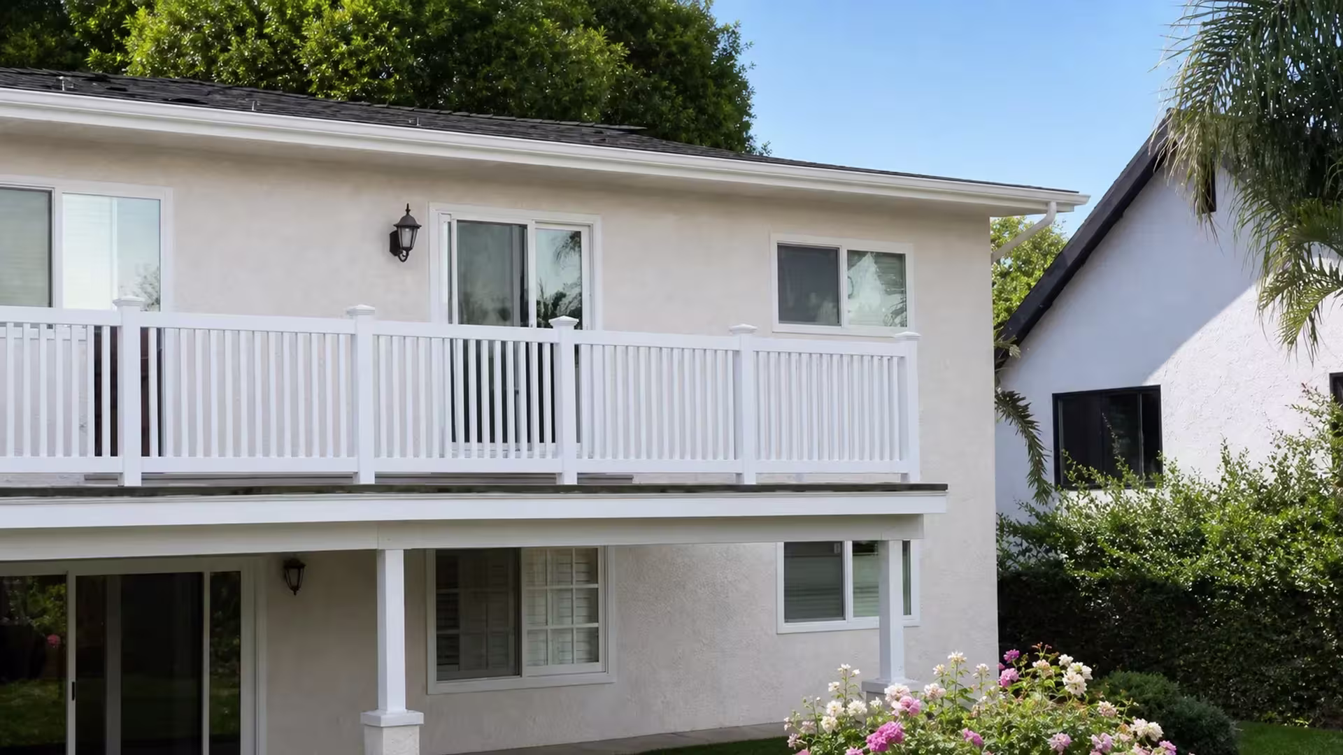 White vinyl balcony railing runs the full length of a second story light stucco house near mature palm trees