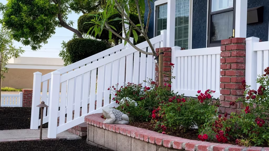 Past a red brick planter wall and rose bed, White vinyl stair railing descends the porch steps