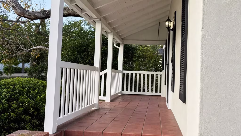 Three connected runs of White vinyl porch railing wrap a covered porch with cream stucco walls and dark shutters