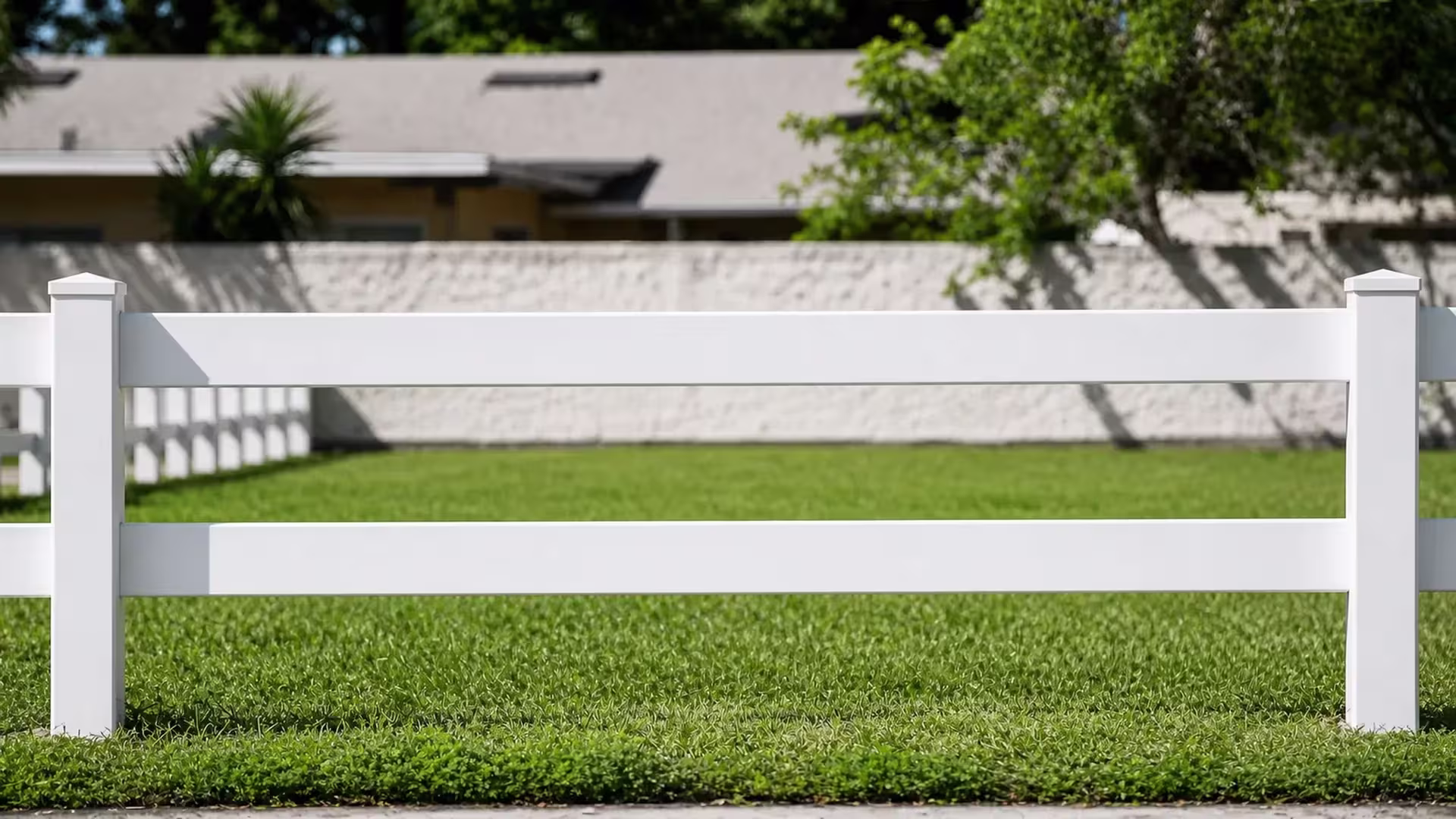 A White vinyl ranch fence with two horizontal rails borders a lawn beside a low concrete wall