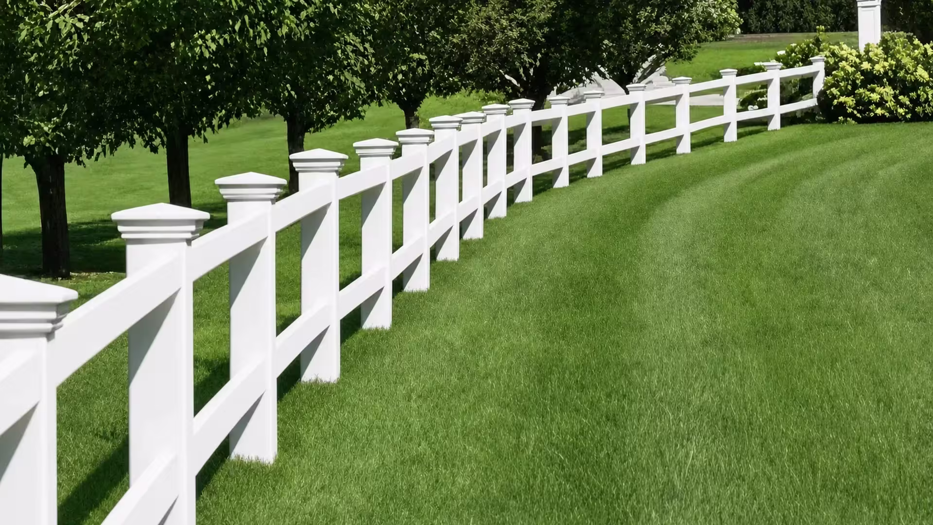 A White vinyl two-rail ranch fence continues past mature trees toward a small structure in the distance