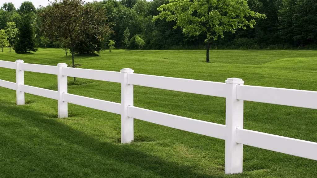 White vinyl ranch fence rails angle across a mown lawn toward a wooded background