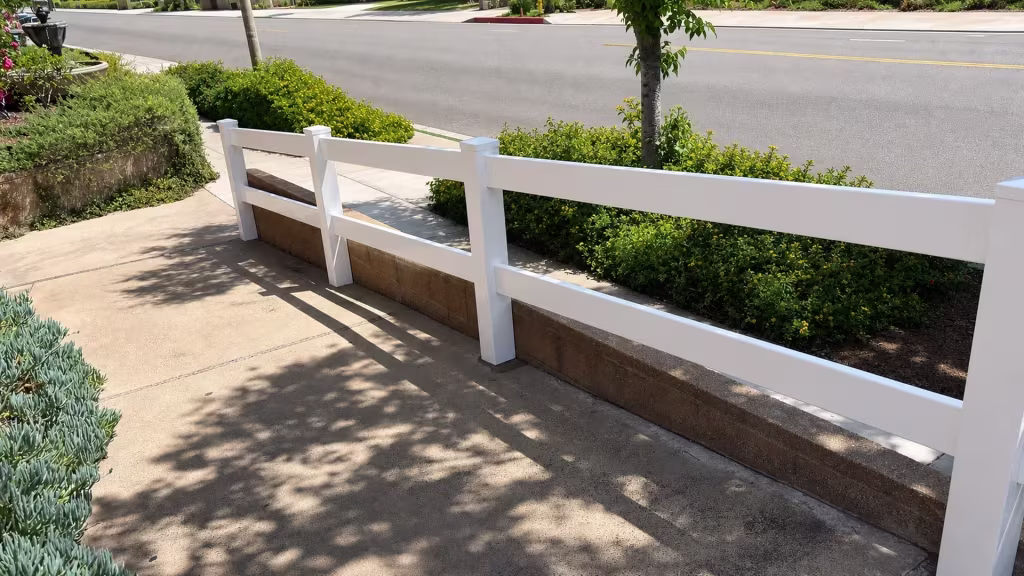 Along a brown masonry retaining wall, White vinyl ranch fencing with two rails traces the inside edge
