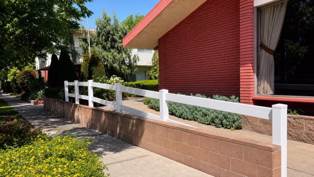 Atop a tan block retaining wall, White vinyl ranch rails run near a stucco residence