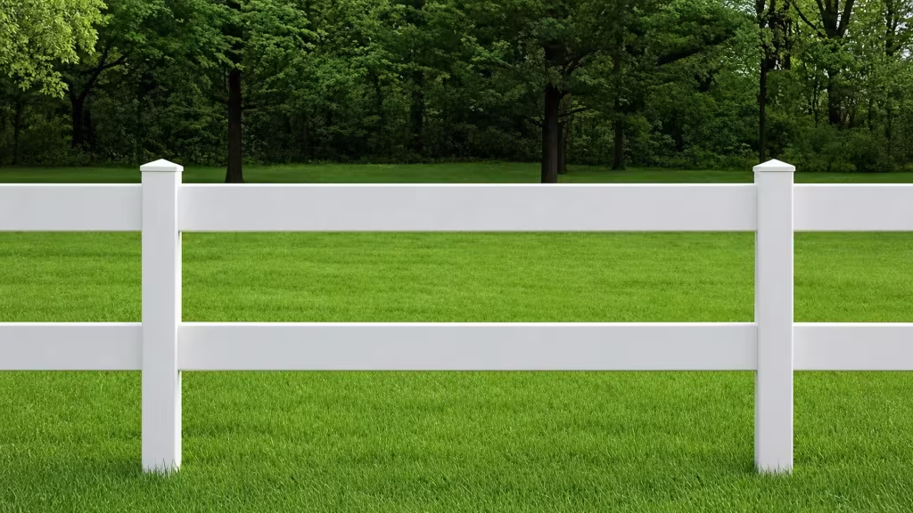 White vinyl ranch rail fencing, two rails high, crosses a lawn in front of deciduous woodland