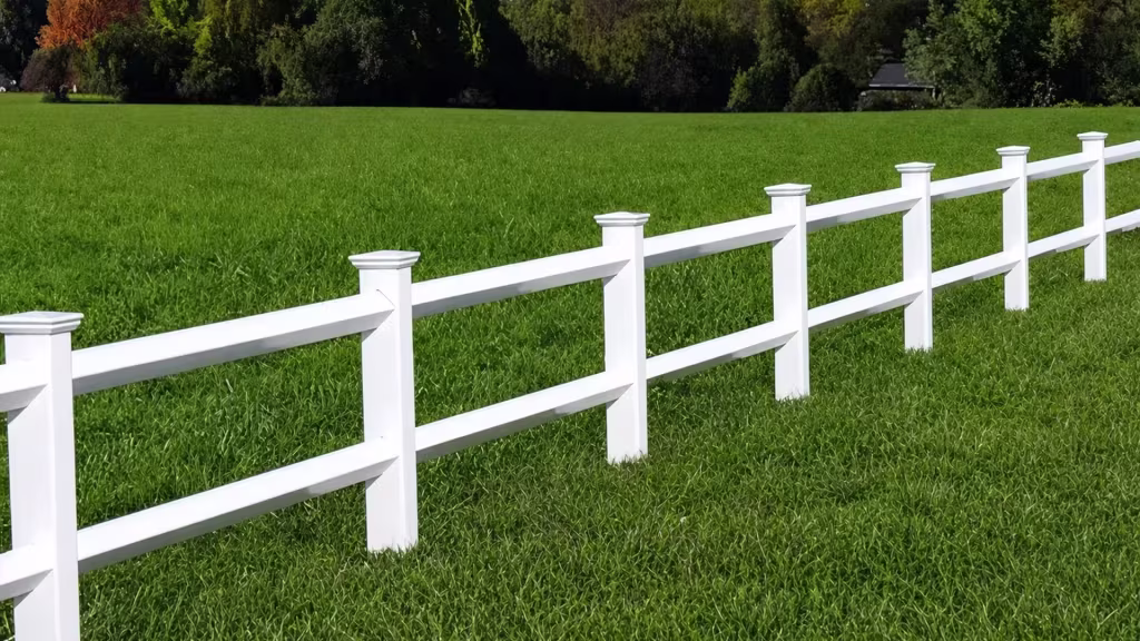 A White two-rail vinyl ranch fence recedes across an open field toward a distant tree line