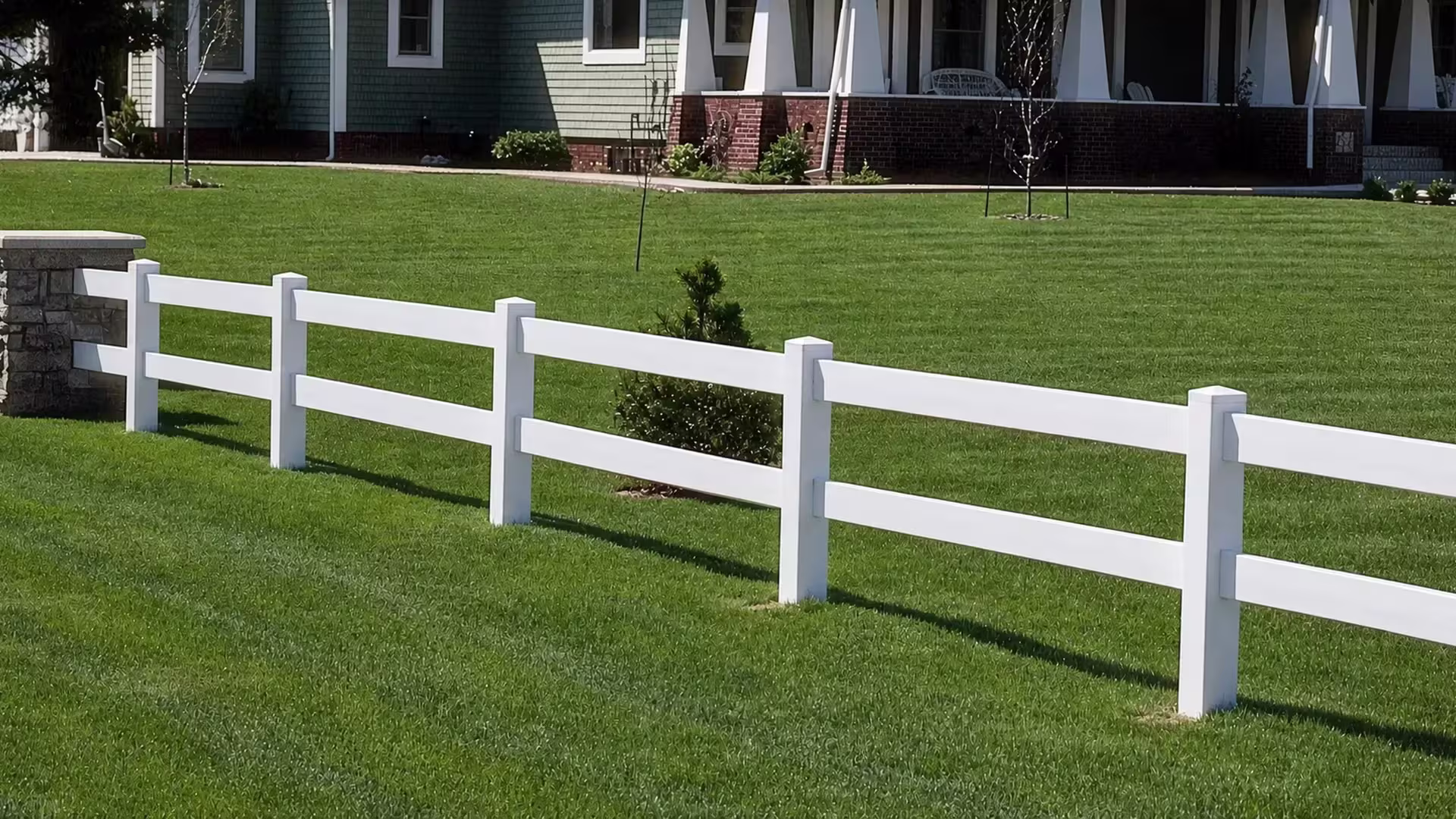 Ranch-style White vinyl fencing with two rails begins at a stone masonry pier near a brick residence