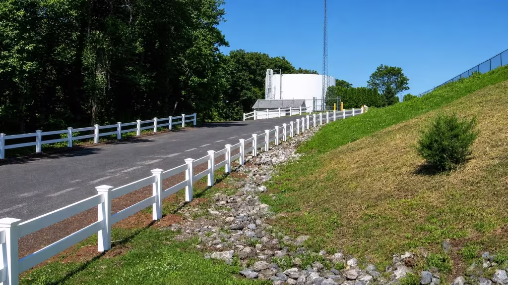 A two-rail White vinyl ranch fence lines both sides of a curving service road near a utility tower