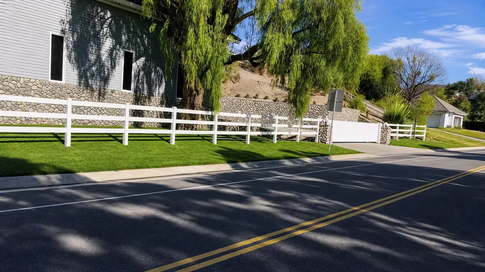 Set on a sloped front lawn, this low White vinyl ranch fence carries three rails beside a stone faced retaining wall
