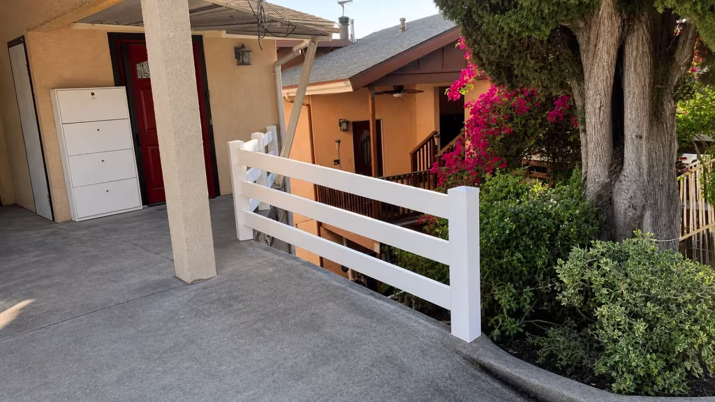 Installed along the edge of a sloped concrete residential walkway, this White vinyl fence carries three ranch rails