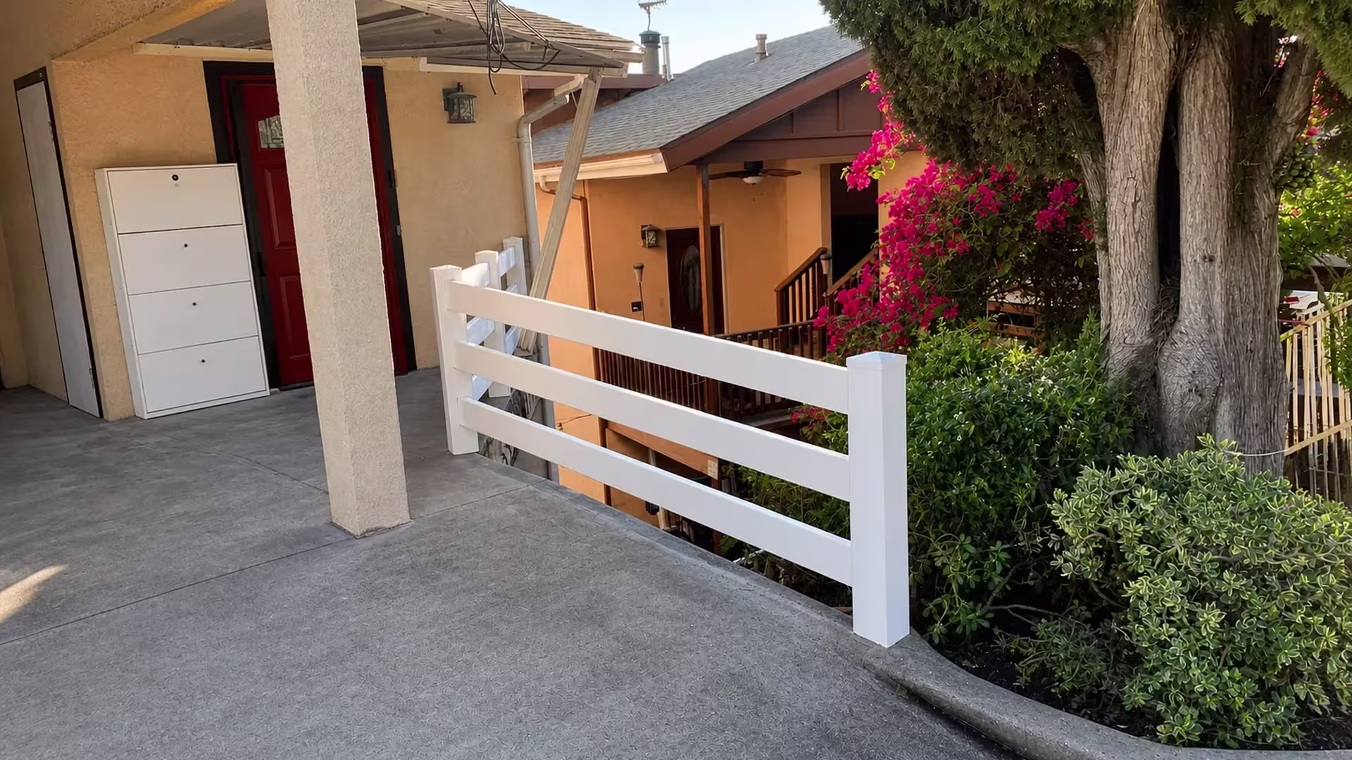 Installed along the edge of a sloped concrete residential walkway, this White vinyl fence carries three ranch rails