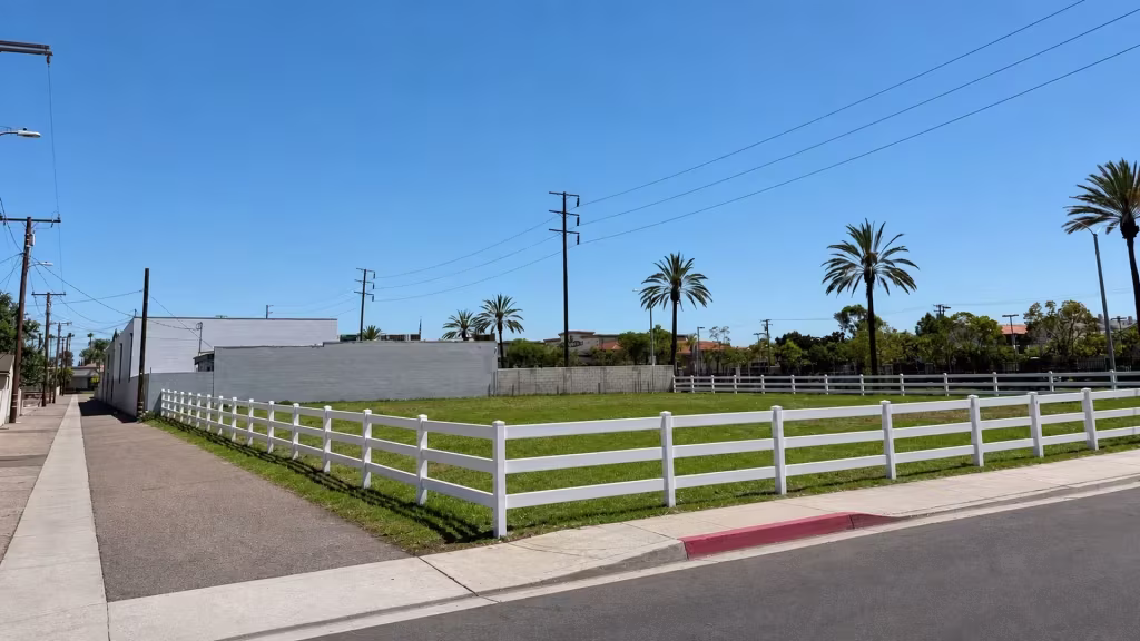 An open corner lot is bordered by White vinyl ranch rail fence set beneath a row of palm trees