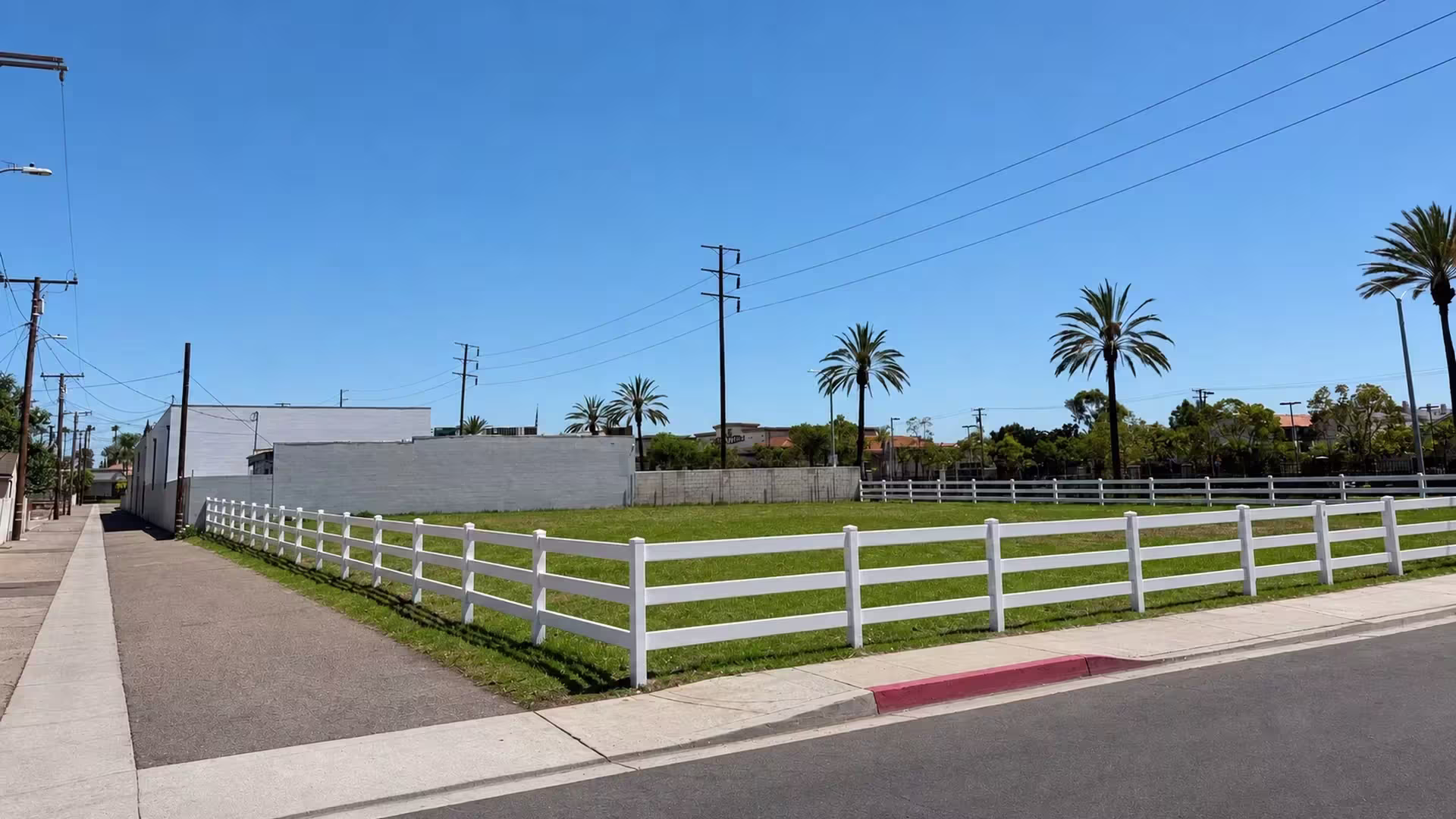 An open corner lot is bordered by White vinyl ranch rail fence set beneath a row of palm trees