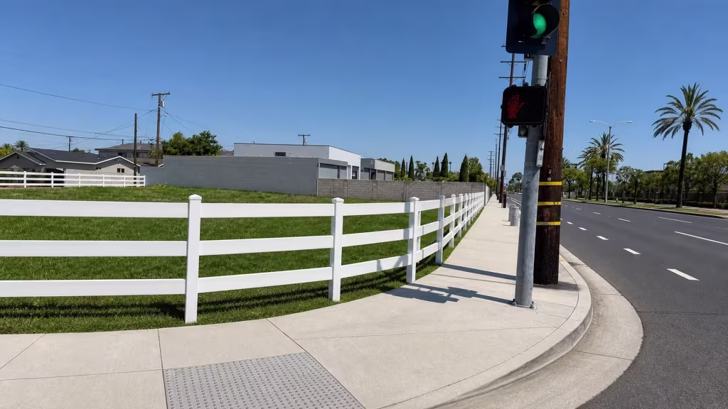 Past low masonry walls and stretches of tactile curb paving, White vinyl ranch rail fence tracks the sidewalk