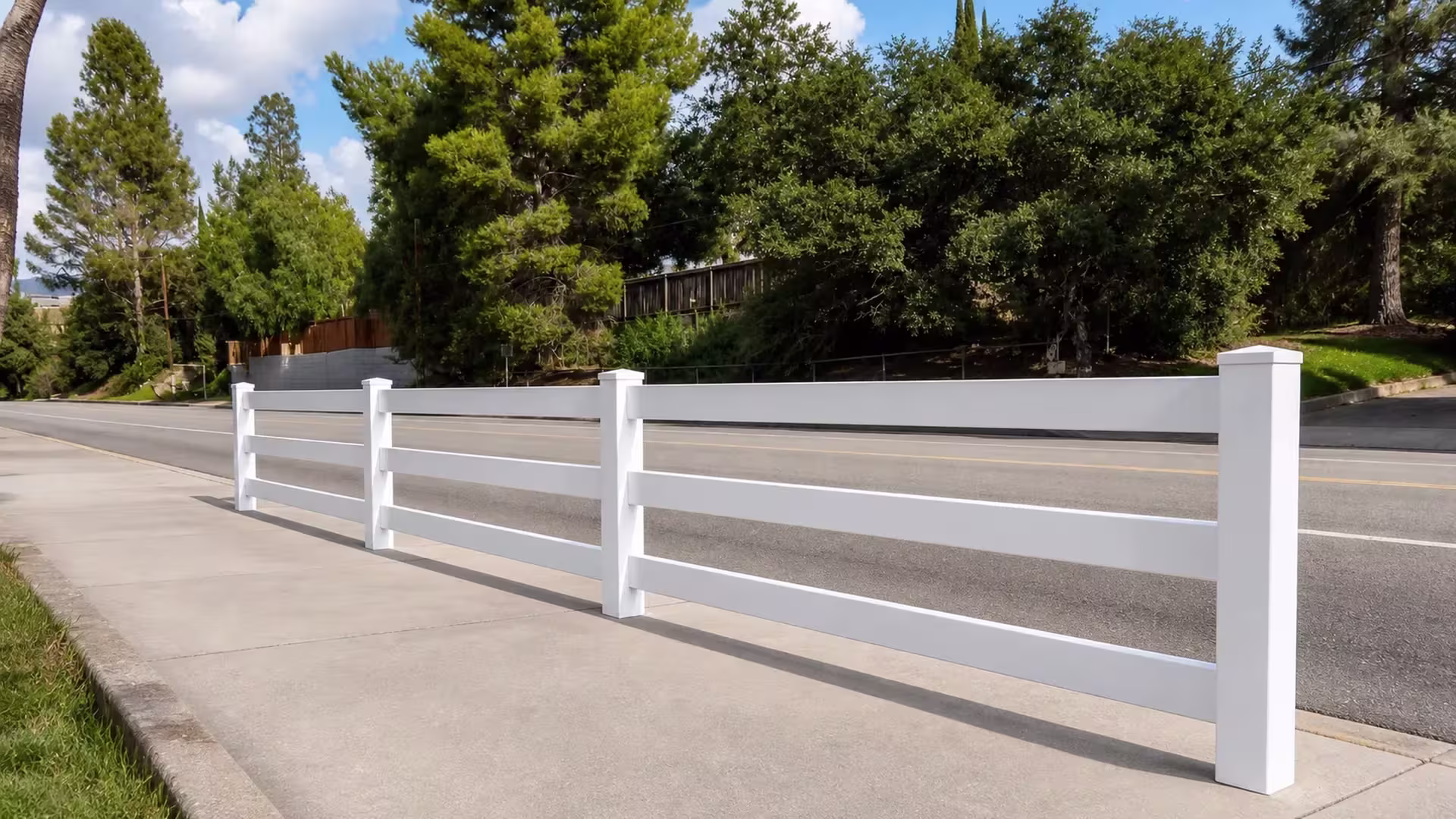 Along the road, a White vinyl ranch rail fence follows the base of a wooded, rising slope lined with mature shrubs