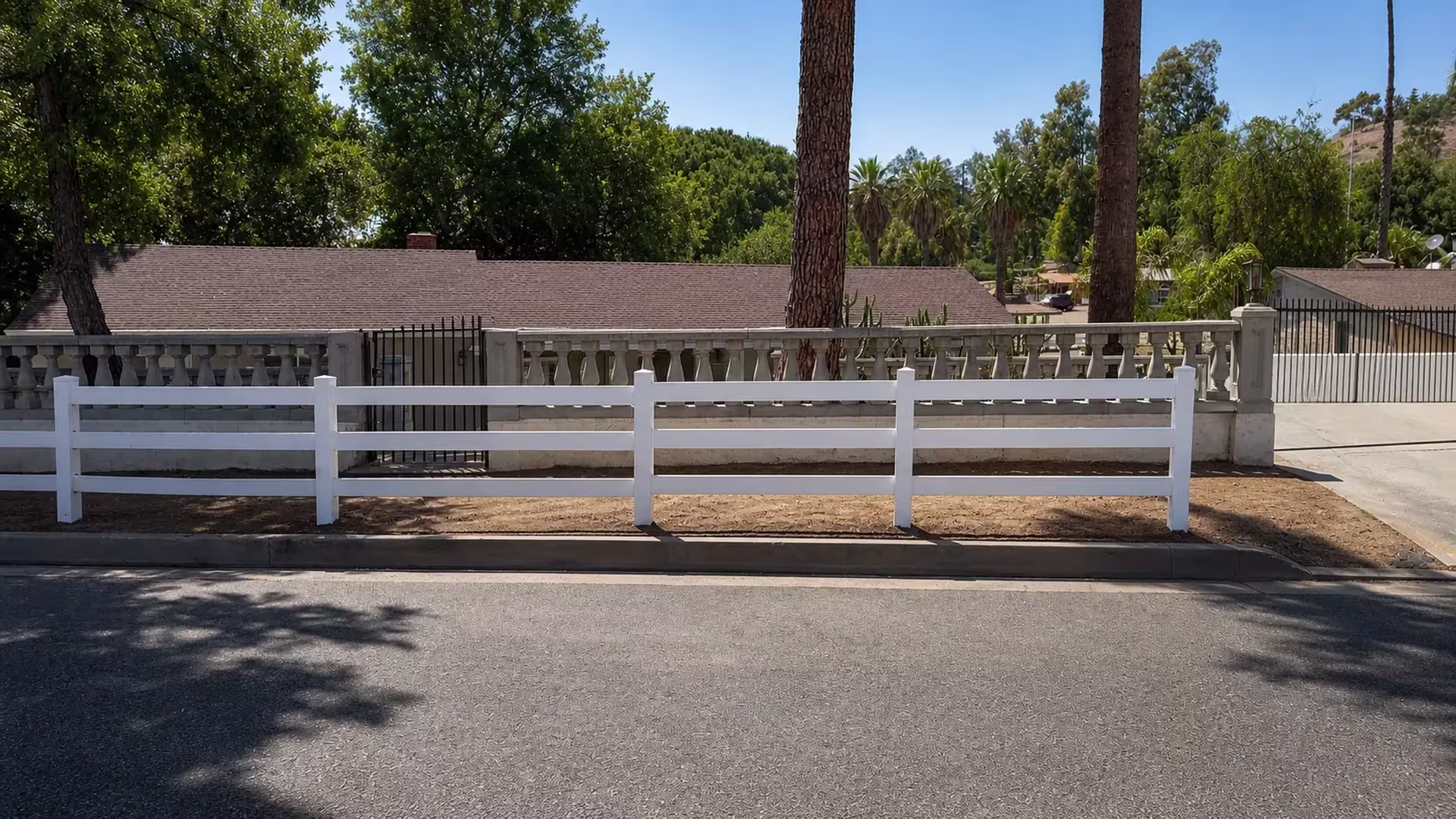 Newly set White vinyl ranch rail fence panels wrap around a mature shade tree near a black metal gate