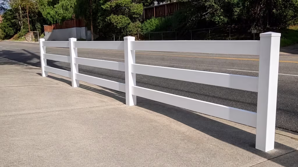 This White vinyl ranch fence, built with three rails, runs along an asphalt street below a hillside retaining wall