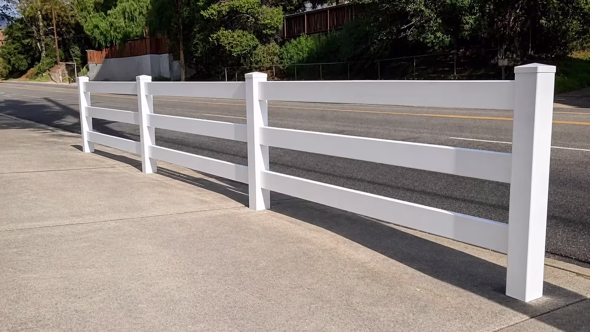 This White vinyl ranch fence, built with three rails, runs along an asphalt street below a hillside retaining wall