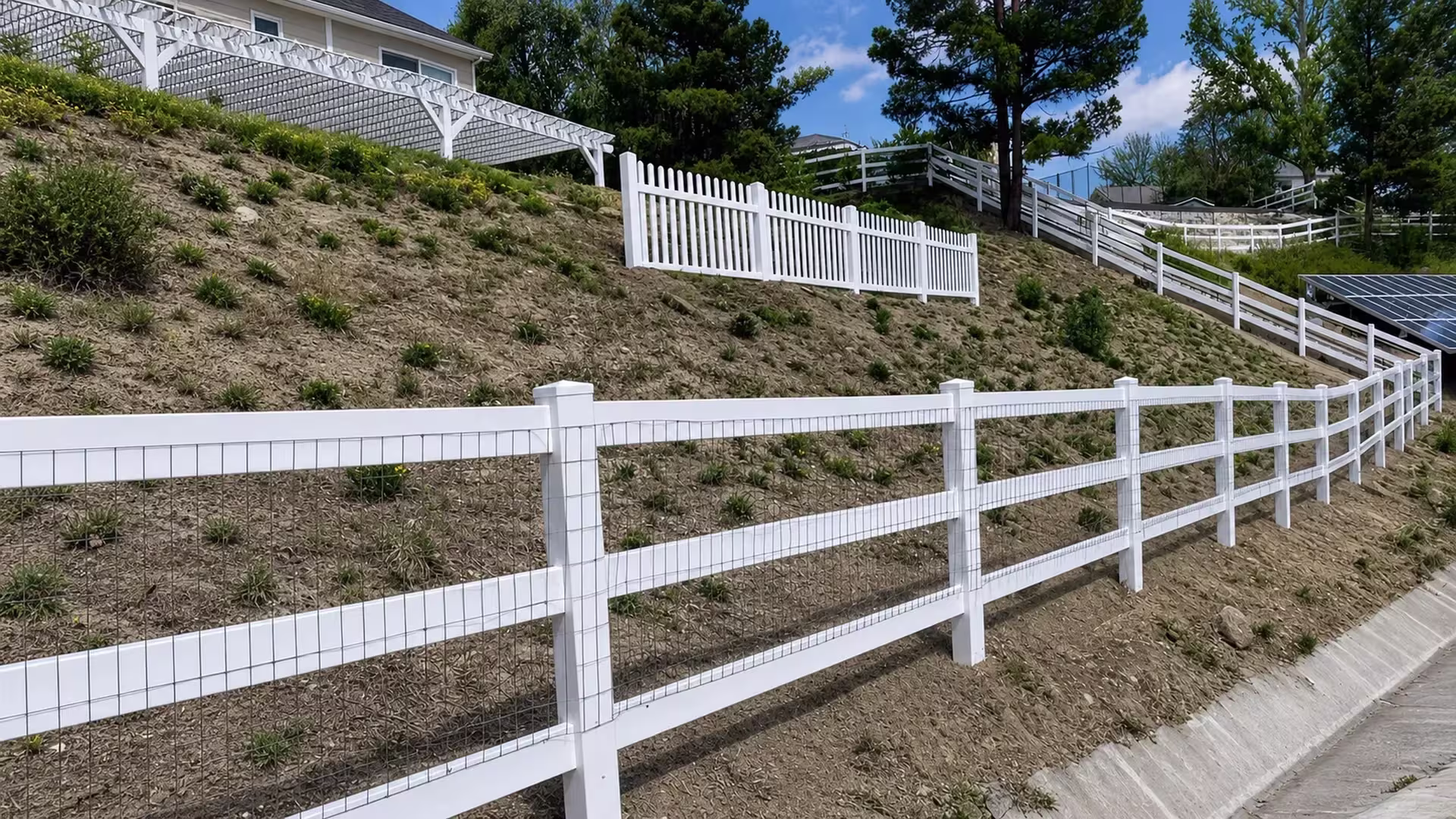Wire mesh backed White vinyl ranch rail fence following a graded hillside slope beside a concrete drainage channel