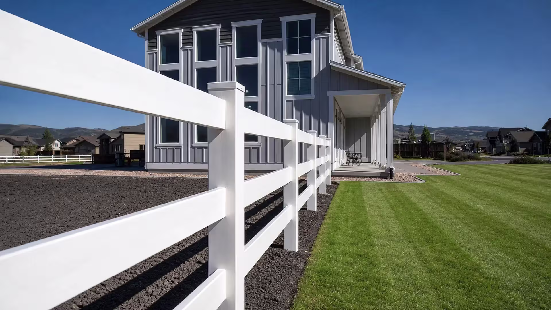 A White vinyl fence with three ranch rails runs diagonally from the foreground toward a gray two story house