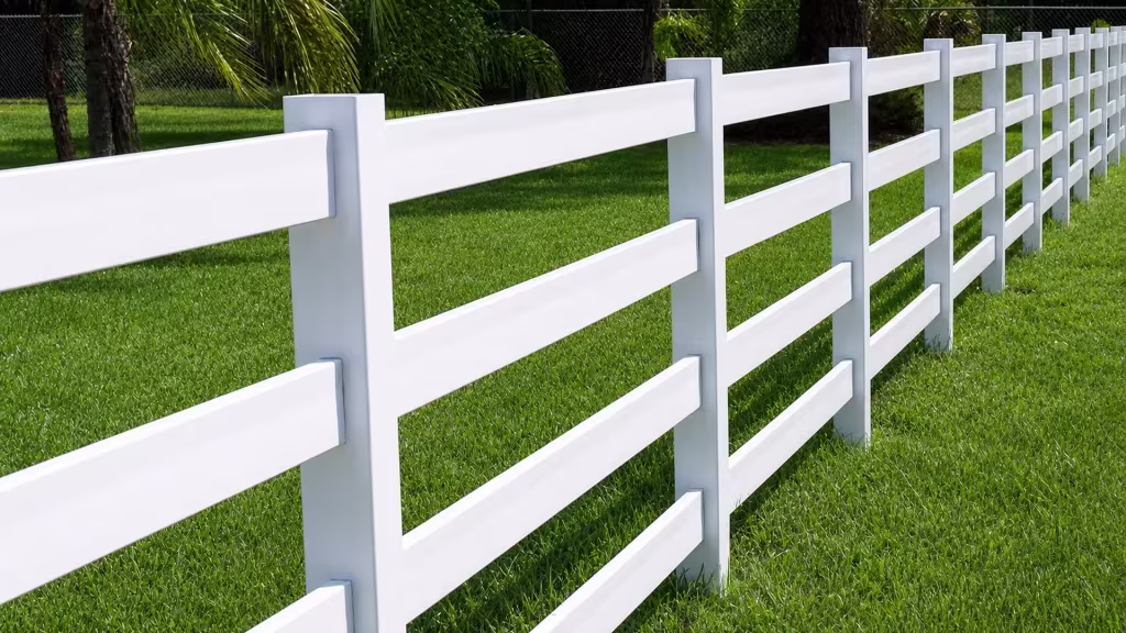 Palm trunks rise over a grass lawn where this White vinyl ranch fence's rails meet a chain-link boundary